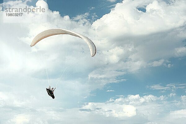 Professioneller Gleitschirmflieger fliegt mit einem weißen Gleitschirm gegen den blaün Himmel und die weißen Wolken. Gleitschirmfliegen Sport