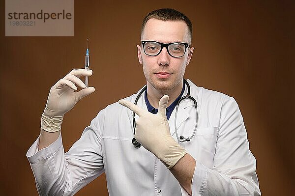 Ein junger kaukasischer Arzt im weißen Kittel und mit Brille hält eine Spritze mit einem Impfstoff. Ein freundlicher  knuddeliger junger Mann zeigt mit dem Finger auf eine Spritze und bietet an  sich impfen zu lassen