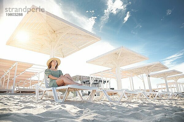 Junge kaukasische Frau im grünen Kleid und Strohhut sitzt auf einer Sonnenliege unter einem Holzschirm an einem weißen Sandstrand am Meer. Raum kopieren. Tourismus und Erholung in heißen Ländern