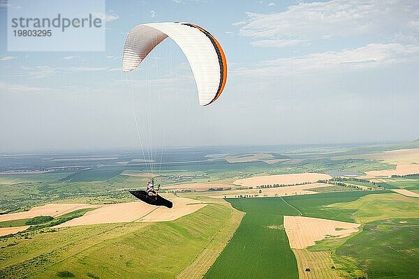 Professioneller Gleitschirmflieger in einem Kokon Anzug fliegt hoch über dem Boden gegen den Himmel und die Felder