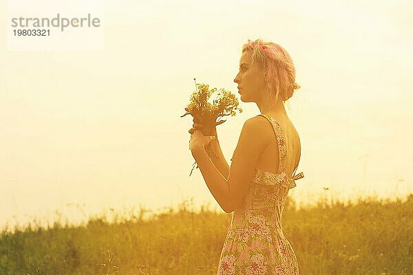 Junge Frau im Kleid mit Blumenstrauß in den Händen bei Sonnenuntergang auf dem Feld. Getöntes warmes Bild