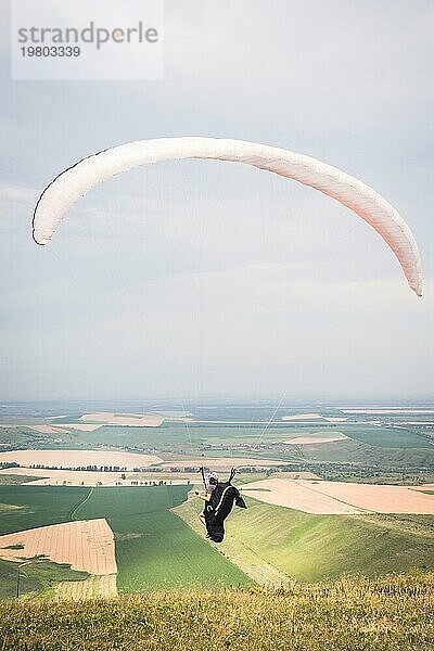 Ein männlicher Gleitschirmflieger  der vom Rand des Berges abhebt  mit Feldern im Hintergrund. Gleitschirmfliegen Sport