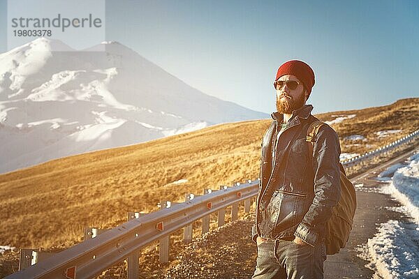 Porträt eines nachdenklichen stilvollen Hipsters mit Bart  Sonnenbrille und Hut mit einem Rucksack vor dem Hintergrund des schneebedeckten Berges Elbrus im Kaukasus bei Sonnenuntergang