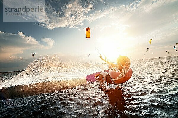 Professionelle Kitesurferin reitet auf einem Brett mit einer Planke in ihren Händen auf einem lemanischen See mit Meerwasser bei Sonnenuntergang. Wasser plätschert und Sonne blendet. Wassersport