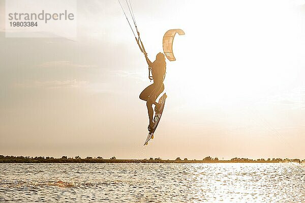 Junge Frau professionelle Kiter führt einen schwierigen Trick in der Luft auf einem schönen Hintergrund des Sonnenuntergangs und Meer