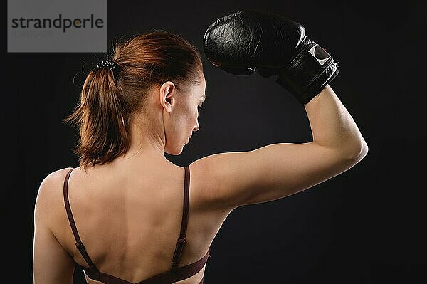Die Rückenansicht einer kaukasischen Frau in Sportkleidung und Boxhandschuhen zeigt eine Geste der Stärke. Zeigt den Bizeps. Studioaufnahme vor einem schwarzen Hintergrund. Frau Kämpferin