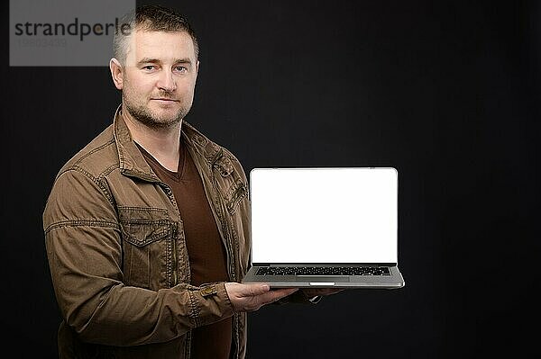 Attraktiver  positiver  freundlicher  kaukasischer Männchen  der einen Laptop in den Händen hält und seinen ausgeschnittenen Bildschirm mit Kopierbereich auf schwarzem Hintergrund präsentiert. Studioaufnahme