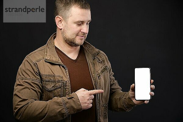 Kaukasischer attraktiver reifer Mann in Freizeitkleidung hält ein Smartphone in den Händen und zeigt mit dem Finger auf den ausgeschnittenen Telefonbildschirm. Ausschnitt des Displays. Studioaufnahme Porträt