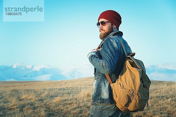 Porträt eines nachdenklichen stilvollen Hipsters mit Bart  Sonnenbrille und Hut mit einem Rucksack vor dem Hintergrund des schneebedeckten Berges Elbrus im Kaukasus bei Sonnenuntergang