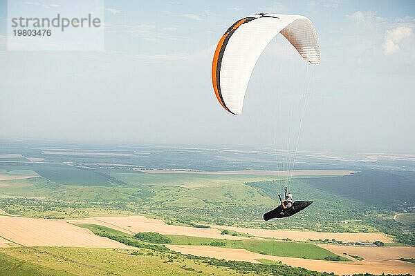 Professioneller Gleitschirmflieger in einem Kokon Anzug fliegt hoch über dem Boden gegen den Himmel und die Felder