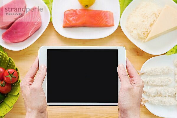 Design Werkstück. Weibliche Hände halten einen Tablet PC über einen Tisch mit rohen Meeresfrüchten  Käse und Gemüse. Blank Display für die Präsentation zum Thema Lebensmittel