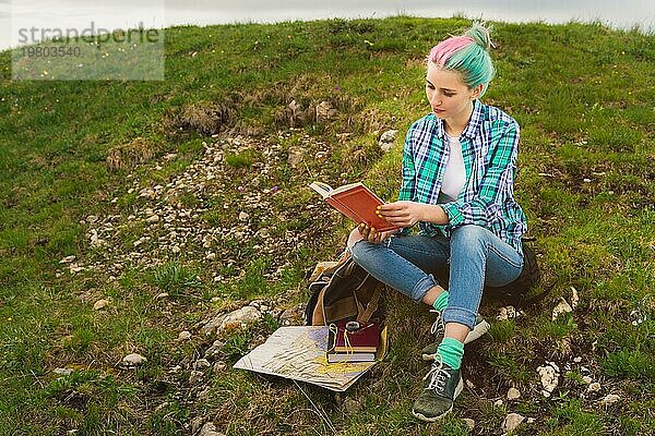 Ein reisendes Mädchen sitzt in den Bergen im Gras und liest ein Buch vor dem Hintergrund epischer Berge. Das Konzept des Lesens während der Erholung und des Urlaubs