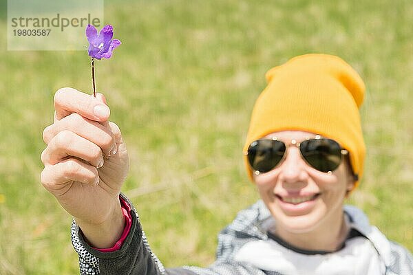 Porträt eines glücklichen  fröhlichen Mädchens mit Hut und Sonnenbrille gibt Ihnen eine Feldblume vor einem Hintergrund von grünen Feldern