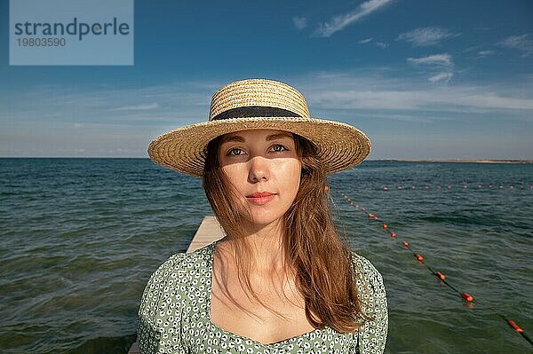 Schönheit am Meer Porträt einer schönen Frau in einem Hut Großaufnahme Attraktives Porträt eines modischen Mädchens mit den Strahlen der Sonne auf ihrem Gesicht vor dem Hintergrund des Meeres und des Himmels