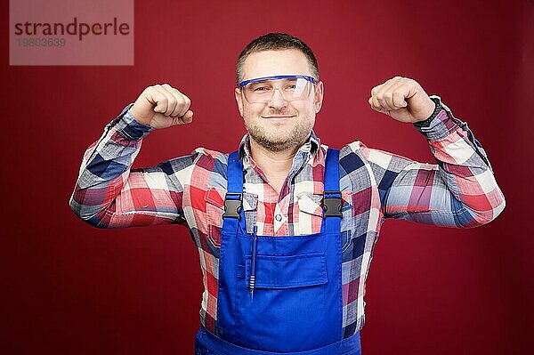 Porträt eines positiven  freundlichen kaukasischen männlichen Vorarbeiters in Latzhose und Schutzbrille auf einem dunkelroten Hintergrund. Er lächelt und schaut in die Kamera. Studioaufnahme. Zeigt eine Geste der Stärke