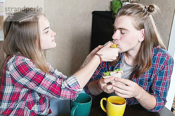 Ein junges  attraktives Paar frühstückt in der Küche ihres Hauses. Sie füttern sich gegenseitig und lachen. Das Konzept der gesunden Ernährung in der Familie und des Glücks
