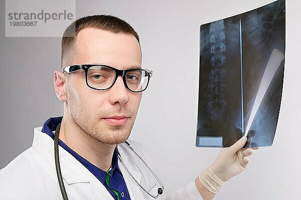 Junger kaukasischer Arzt und Radiologe hält ein Röntgenbild in der Hand und schaut in die Kamera. Ein Mann mit Brille und Uniform  der ein Stethoskop um den Hals trägt. Studioaufnahme auf grauem Hintergrund