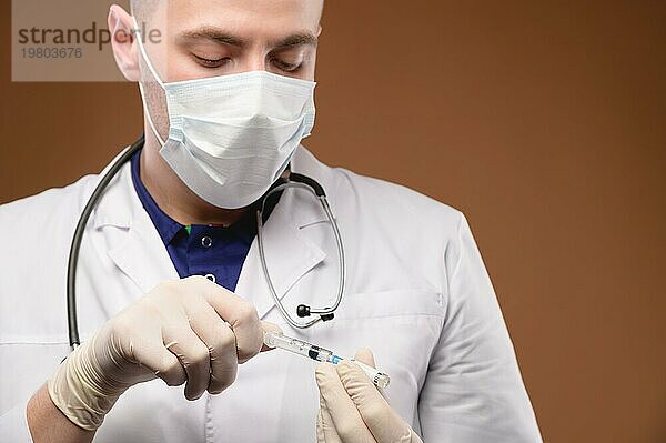 Ein junger weißer Arzt in Kittel und Maske zieht einen Impfstoff aus einer Ampulle in eine Spritze. Fokus auf die Spritze