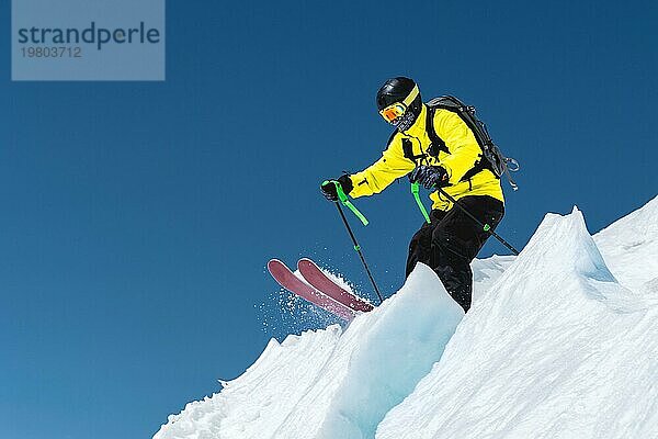 Ein Skifahrer in voller Sportausrüstung springt von der Spitze des Gletschers in den Abgrund vor dem Hintergrund des blaün Himmels und der schneebedeckten Berge des Kaukasus. Elbrus Gebiet. Russland