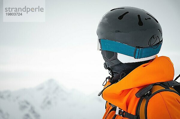 Nahaufnahme Ein professioneller Skifahrer in roter Jacke  Helm und Maske steht auf einer Piste vor der Kulisse eines Gletschers und epischer Berge und zeigt mit einem Skistock auf eine Bergkette Freeride Konzept