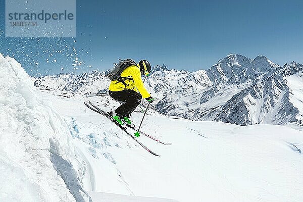 Ein Skifahrer in voller Sportausrüstung springt von der Spitze des Gletschers in den Abgrund vor dem Hintergrund des blaün Himmels und der schneebedeckten Berge des Kaukasus. Elbrus Gebiet. Russland