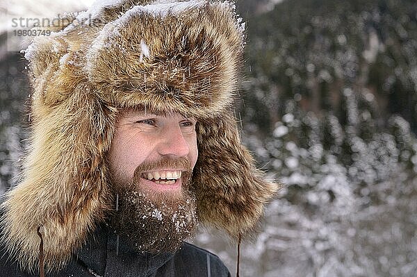 Mann in einem Pelz Wintermütze mit Ohren lächelnd Porträt auf einem Hintergrund von einem Winterwald