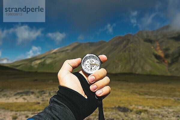 Blickpunktaufnahme. Eine Ichperspektive eines Mannes  der einen Kompass vor dem Hintergrund einer epischen Landschaft mit Klippen  Hügeln und einem blaün Himmel mit Wolken hält