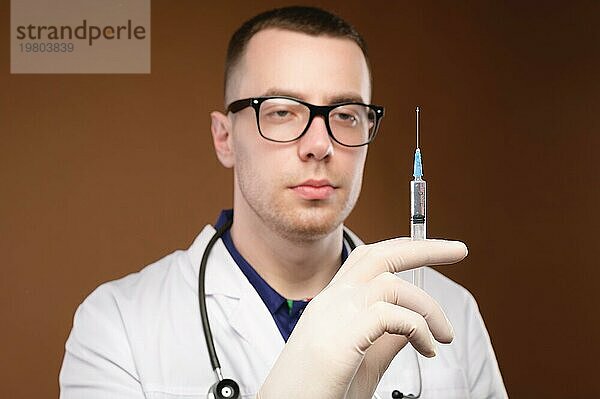 Kaukasischer Arzt im Bademantel mit Stethoskop und Handschuhen  der eine Spritze hält  vor einem schlichten Hintergrund  nachdenklicher Blick  Studioaufnahme