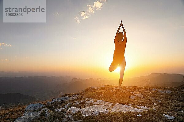 Silhouette einer Frau in einer Baumpose Yogaasana steht in der Nähe einer Klippe in den Bergen vor dem Hintergrund eines Sonnenuntergangs mit einem klaren Himmel