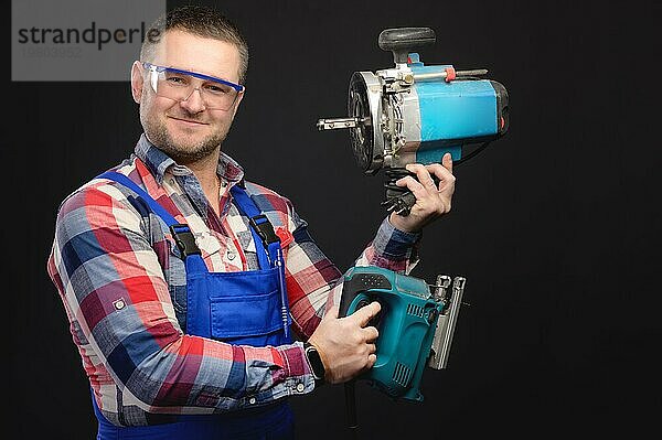 Porträt eines lächelnden jungen Baumeisters  der Werkzeuge in beiden Händen hält  während er steht und in die Kamera schaut  auf schwarzem Hintergrund