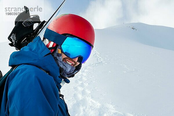 Ausgerüsteter Skifahrer  der die Skier auf der Schulter hält und direkt in die Kamera schaut  Porträt. Mann mit Winterausrüstung in den Bergen in einem Skigebiet