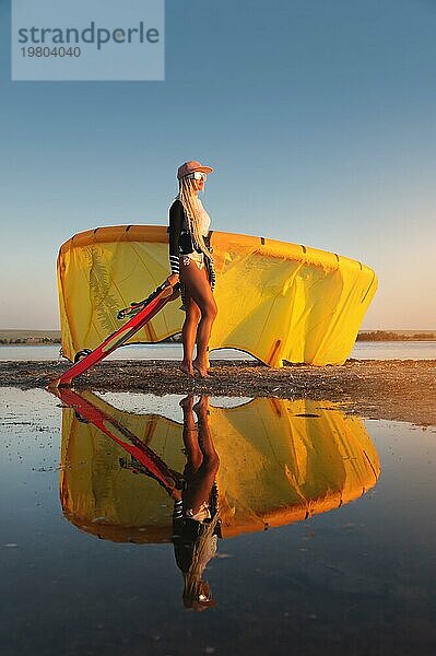 Ein junges sexy Mädchen in einem Neoprenanzug und Sonnenbrille steht mit einer gelben Seeschlange bei Sonnenuntergang an der Küste eines Strandes im Sand