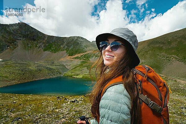 Schöne junge Frau mit einem Touristenrucksack in einer Sportjacke und Hut. Sommerlandschaft von See in grünen Bergen