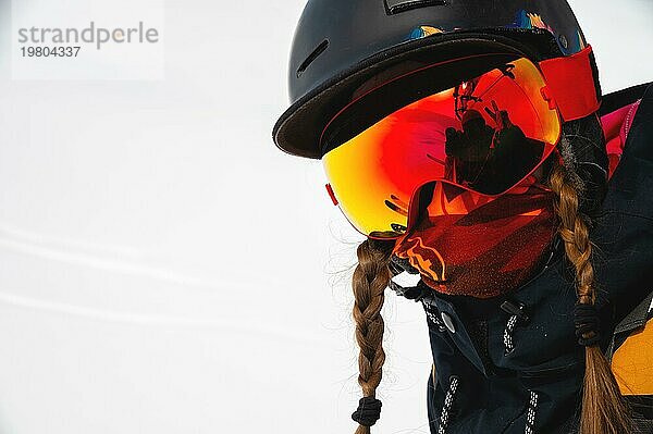 Porträt einer jungen Frau in einem Skigebiet vor der Kulisse eines schneebedeckten Berges  ihre Freunde sind in einer Maske dargestellt  ein fröhlicher Urlaub. Wintersport