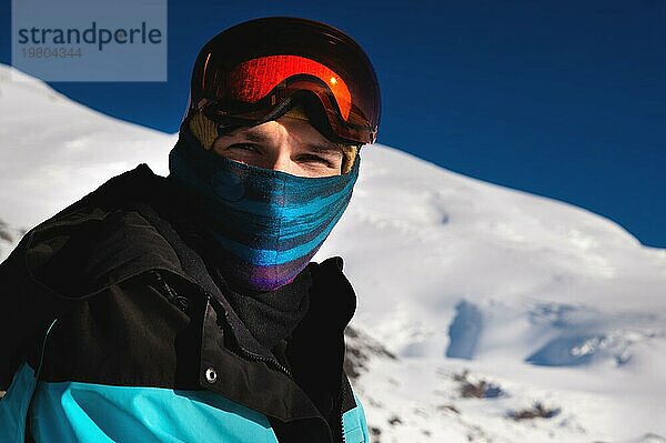 Porträt eines Skifahrers in den Bergen ohne Skimaske mit einem Schal im Gesicht  der mit zusammengekniffenen Augen in die Kamera blickt  vor dem Hintergrund der schneebedeckten Berge