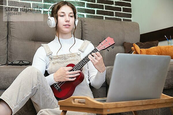 Eine junge  hübsche Frau sitzt zu Hause auf dem Boden und lernt Gitarre oder Ukulele spielen  neben einem Laptop und einer Tasse Kaffee