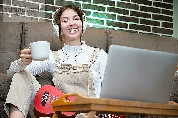 Fröhliche junge Frau sitzt mit einer Tasse Kaffee und einer Ukulele  trägt Kopfhörer auf dem Boden im Wohnzimmer ihres Hauses und schaut in den LaptopMonitor Fernunterricht