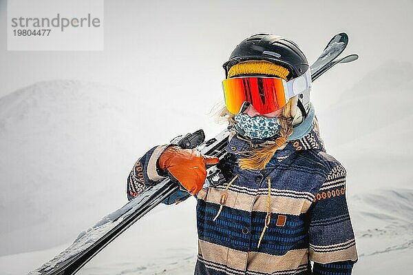 Porträt einer hübschen und aktiven Skifahrerin  die eine Maske trägt und Skier in den Händen hält  aktiver Winterurlaub. Extrem Lifestyle Konzept