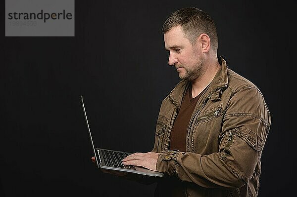 Porträt eines optimistischen  seriösen Mannes im Hemd  der mit einem Laptop in der Hand steht  auf das Display schaut und arbeitet. Studioaufnahme auf schwarzem Hintergrund  Porträt