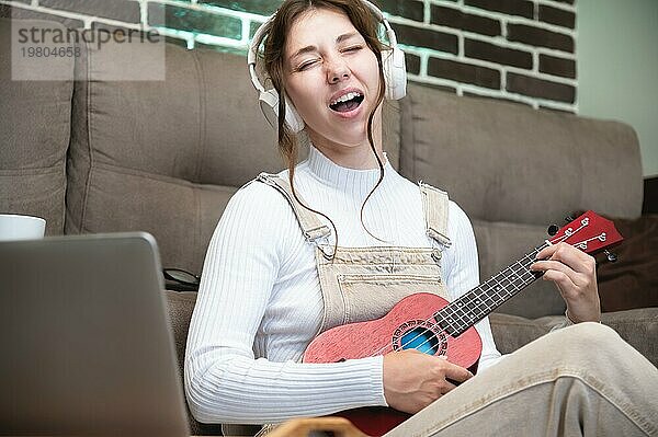 Junge Studentin  die zu Hause auf dem Boden im Wohnzimmer vor einem Laptop sitzend Gesang lernt und Ukulele spielt