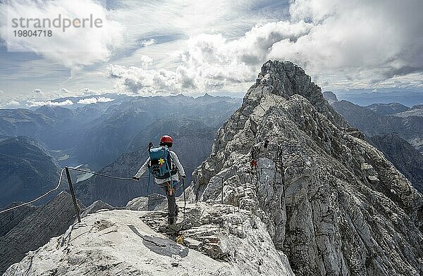 Bergsteigerin in einem mit Stahlseil gesicherten Klettersteig  schmaler felsiger Berggrat  Watzmann Überschreitung zur Watzmann Mittelspitze  Ausblick auf Bergpanorama mit Steinernes Meer  Nationalpark Berchtesgaden  Berchtesgadener Alpen  Bayern  Deutschland  Europa