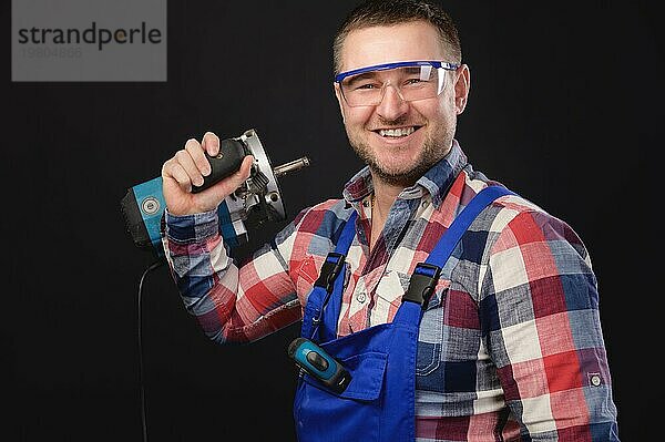 Ein lächelnder Mann in einem Schutzanzug und einem karierten Hemd  der eine Schutzbrille trägt  steht und hält ein Reparaturwerkzeug in den Händen. Porträt auf schwarzem Hintergrund