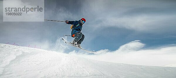 Ein Sportler Skifahrer in Skiausrüstung springt einen steilen verschneiten Hang eines Berges vor dem Hintergrund eines blaün Himmels und schneebedeckten Bergen hinunter. Winter riskante Sportarten  Mut und Geschwindigkeit Konzept