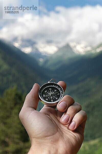 Magnetischer Kompass in der Handfläche eines Mannes vor dem Hintergrund einer Bergkette in den Wolken im Sommer im Freien  Reisen  Ichperspektive