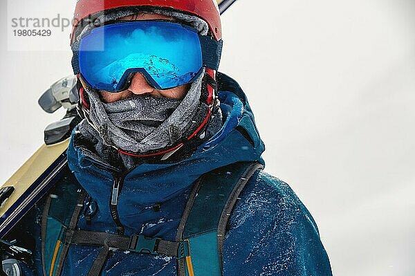 Ausgerüsteter Skifahrer  der die Skier auf der Schulter hält und direkt in die Kamera schaut  Porträt. Mann mit Winterausrüstung in den Bergen in einem Skigebiet