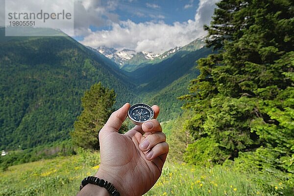 Ein Mann hält einen Kompass in der Hand vor der Kulisse der Natur  einem grünen Wald und Bergen in flauschigen Wolken