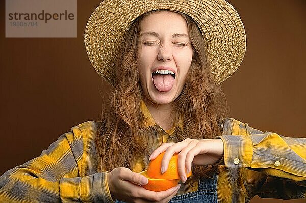 Attraktive kaukasische junge Frau in einem karierten Hemd und einem Strohhut mit einer aufgeschnittenen Orangenfrucht in den Händen schreit und schneidet Grimassen wegen des sauren Geschmacks