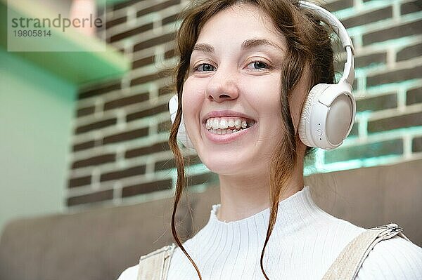 Porträt einer lächelnden jungen kaukasischen Frau mit Kopfhörern im Gespräch über Videoanruf von zu Hause aus. Foto einer glücklichen Frau mit Headset  die in die Kamera schaut und ein virtuelles Gespräch oder eine Webcam Konferenz führt