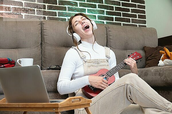 Musikunterricht aus der Ferne  Ukulele spielen oder Gesangsunterricht  Singen. Hipster Schüler  der eine Unterrichtsstunde aufnimmt oder mit einer Laptop Webcam lernt  während er auf dem Boden im Wohnzimmer sitzt