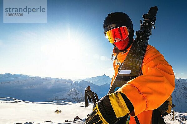 Foto eines Sportlers mit Helm und Maske und Skiern auf der Schulter. Ein junger Mann steht in den Bergen vor dem Hintergrund der Sonne und schaut in die Kamera  während er die Skier hält